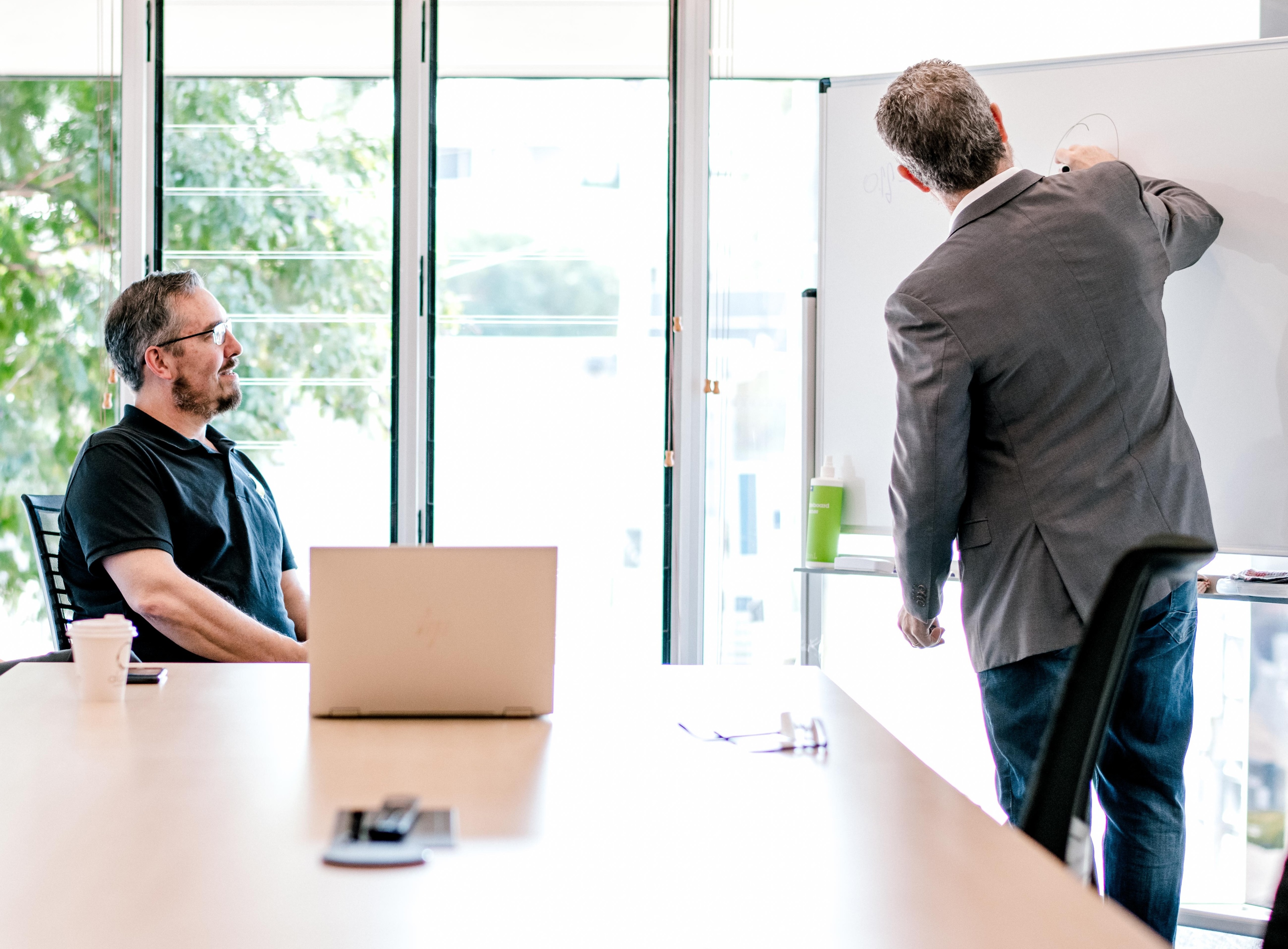 A man in a jacket stands up and writes on a whiteboard while another man sits at the table and watches.