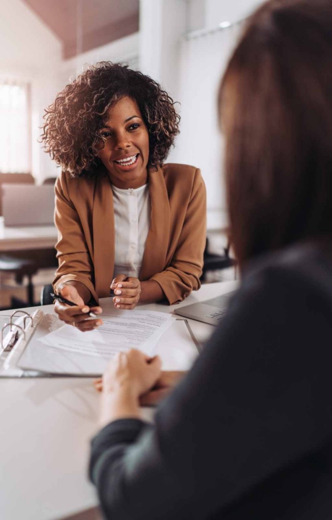 Meeting between two women. Woman with dark curly hair, mustard coloured jacket and white shirt is on focus on the other side of the desk.