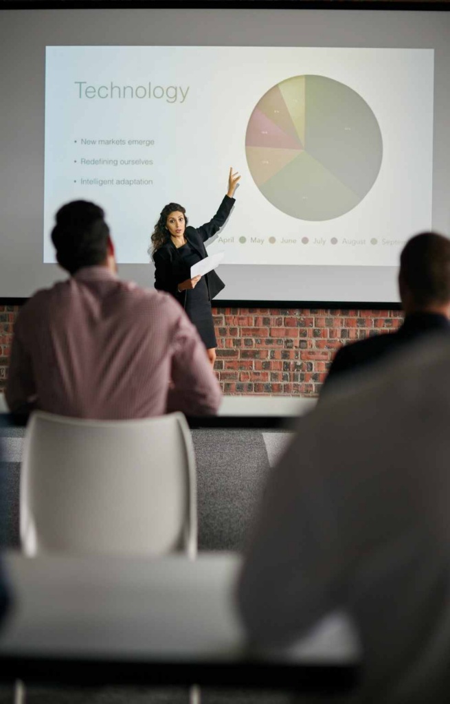 Woman giving a presentation to an audience. She is standing up and pointing to the slides being projected behind her on the wall.